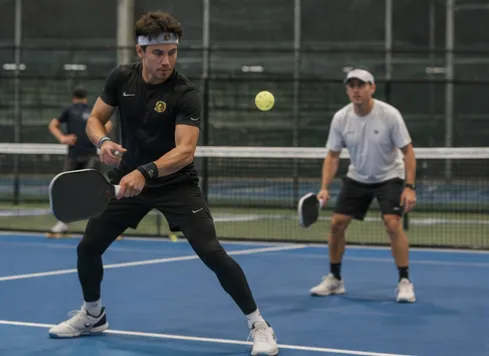 An athletic player in black compression gear striking a forehand with intense focus, another player visible across the court.
